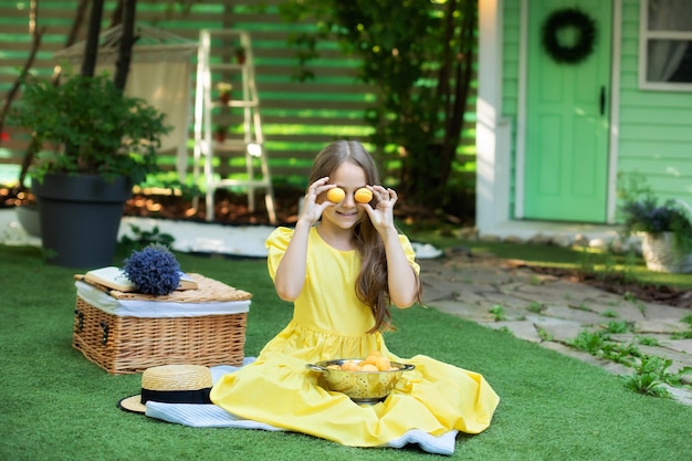 Smiling Little girl in yellow dress indulges with fruit on green lawn at summer.