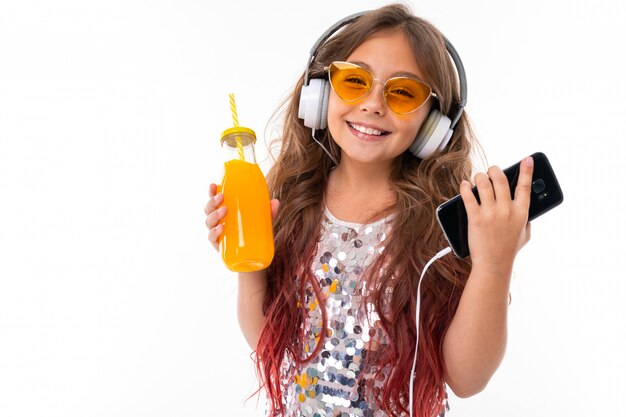 Smiling little woman in headphones listenes to music in headphones at white surface wall