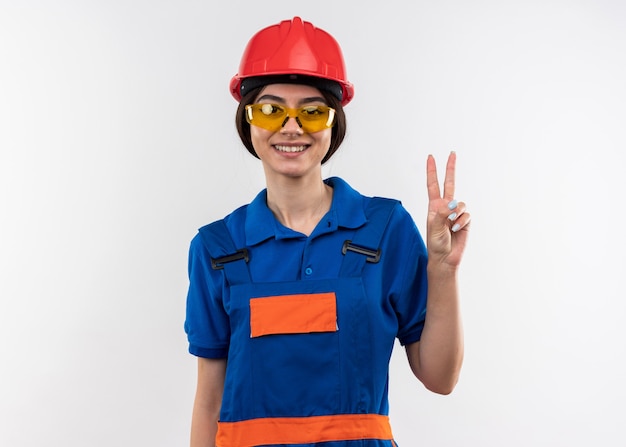 Smiling looking at camera young builder woman in uniform with glasses showing peace gesture 