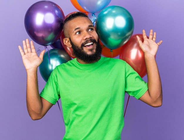 Smiling looking side young afro-american guy wearing green t-shirt standing in front balloons spreading hands isolated on blue wall