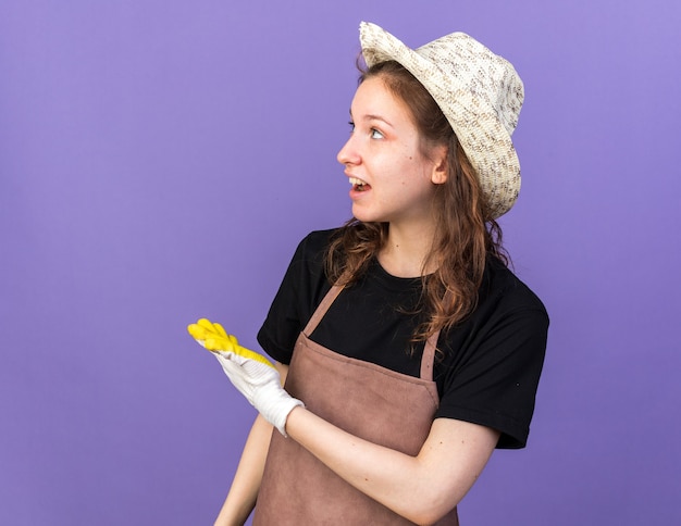 Smiling looking side young female gardener wearing gardening hat with gloves points at behind 