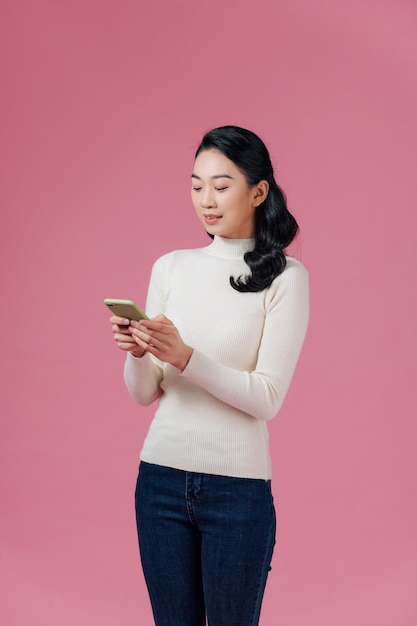 Smiling lovely young woman standing and using cell phone over pink background