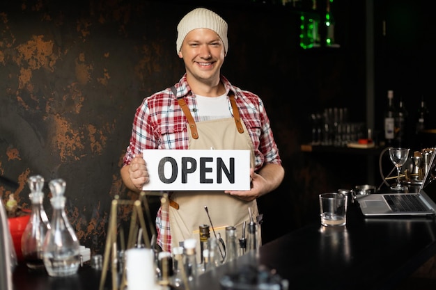 Smiling male bar owner stands behind the bar and holds the sign open He invites everyone to enter Open