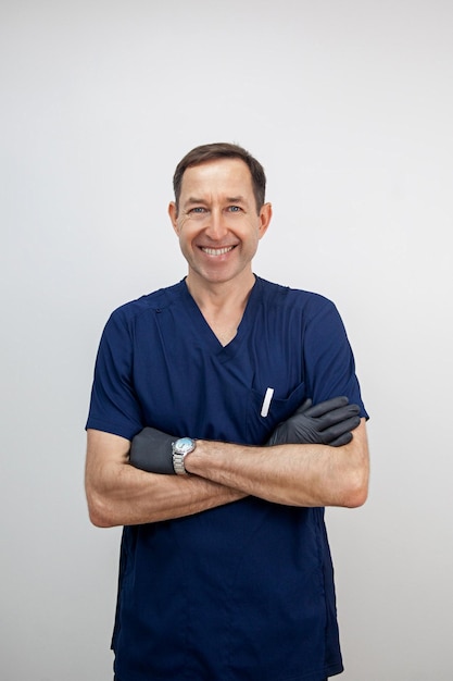 Smiling male cosmetologist doctor with crossed arms in a blue medical uniform and black gloves White background