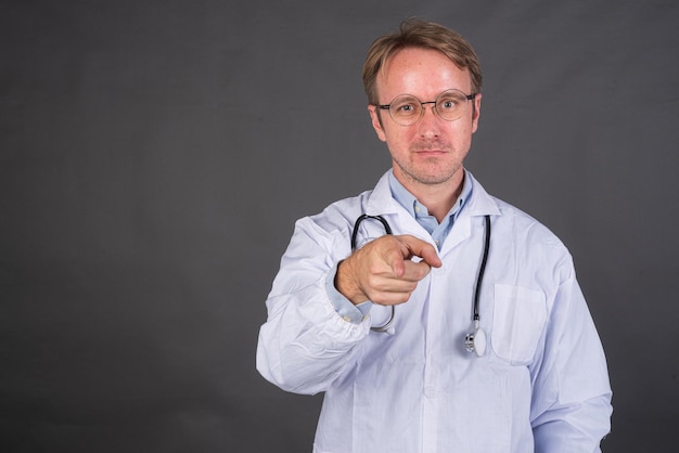 Smiling male doctor with stethoscope in medical coat pointing finger against gray background