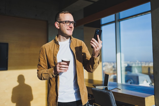 Smiling male worker in casual clothes looking away while talking on mobile phone and drinking coffee during break in modern creative workspace