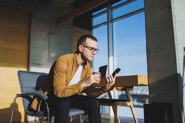 Smiling male worker in casual clothes looking away while talking on mobile phone and drinking coffee during break in modern creative workspace