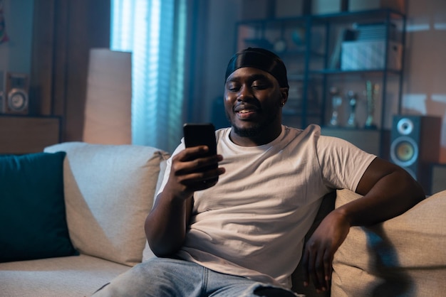 Smiling man in black kerchief on head with dark skin is resting in evening on couch in dark room