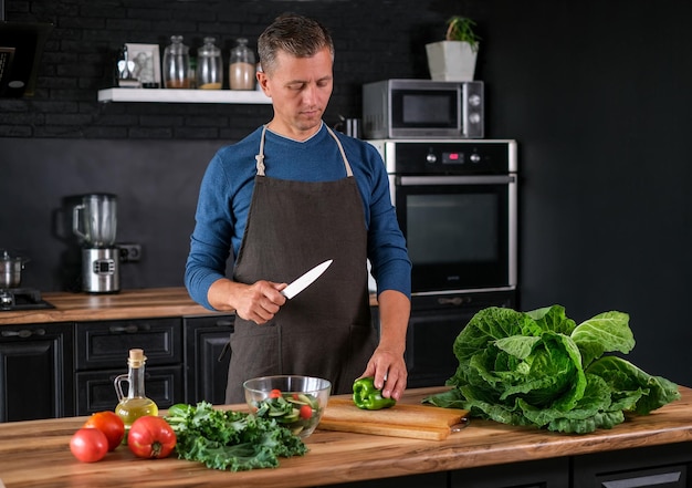 Smiling  man cooking salad, cutting fresh vegetables in modern black kitchen