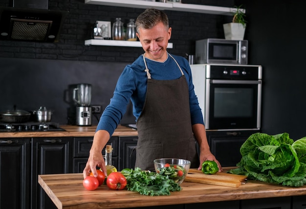 Smiling  man cooking salad, cutting fresh vegetables in modern black kitchen