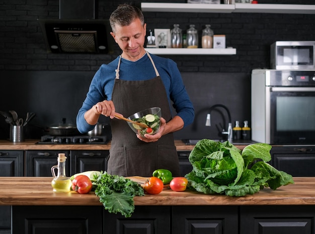 Smiling  man cooking salad, cutting fresh vegetables in modern black kitchen