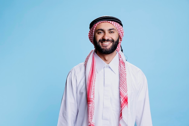 Smiling man dressed in traditional muslim clothes standing with cheerful expression studio portrait Happy arab person posing in thobe and headscarf while looking at camera with carefree emotion