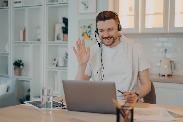 Smiling man freelancer in headphones waving to his colleague and having online meeting, working remotely from home, happy guy looking at laptop webcam and greeting somebody. Freelance concept