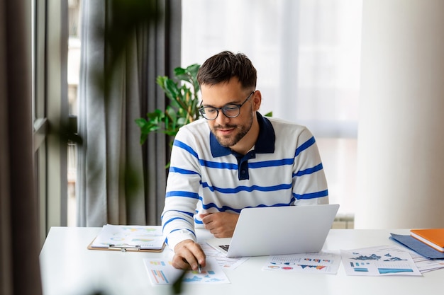 Smiling man in glasses sit at desk in office looking at data while working on his laptop