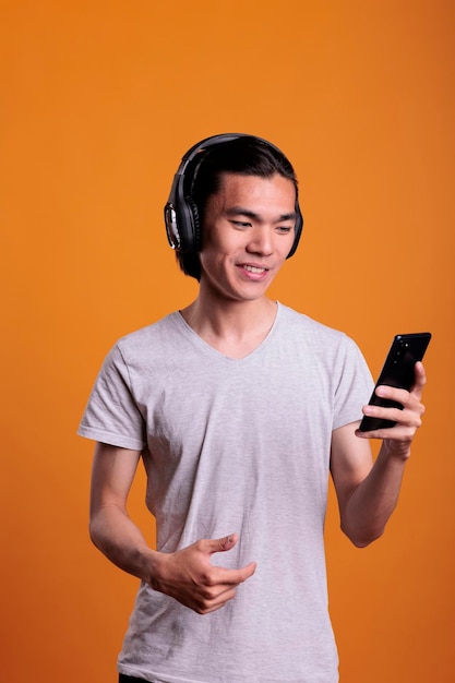 Smiling man in headphones watching funny video on mobile phone, asian teenager listening to podcast. Young adult person wearing wireless earphones, enjoying music on smartphone