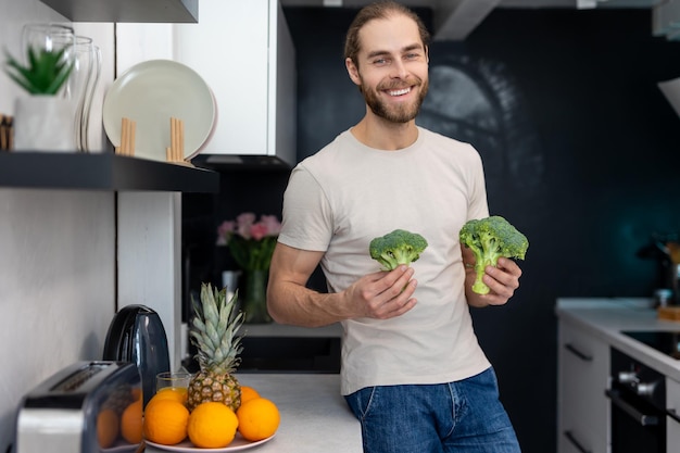 Smiling man holding green fresh broccoli standing in modern kitchen at home