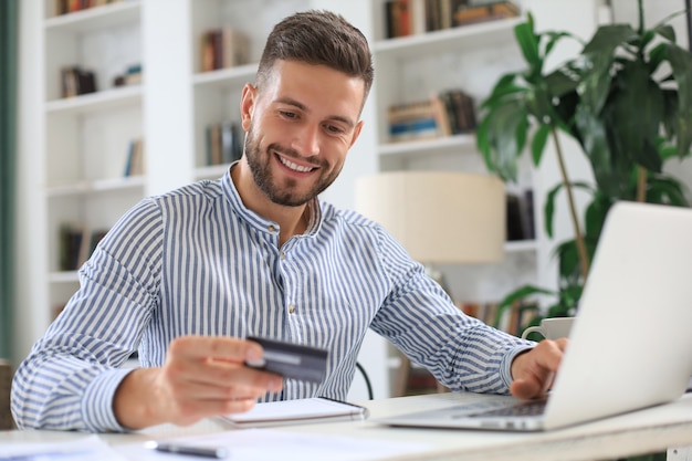 Smiling man sitting in office and pays by credit card with his laptop.