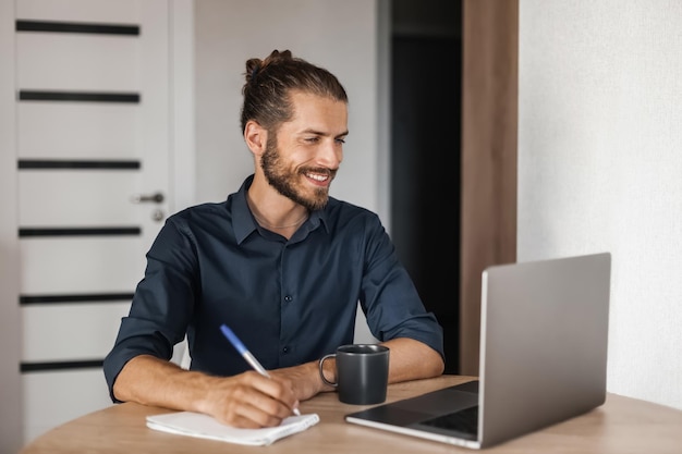 Smiling man sitting at a table and writing in a notebook while looking into a laptop