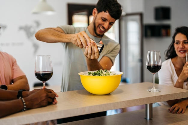 Smiling man sprinkling salt on a salad among friends drinking red wine