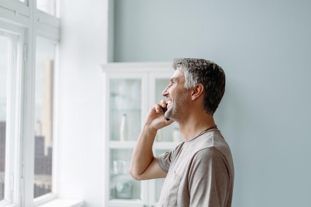 Smiling man talking on a smartphone standing near the window