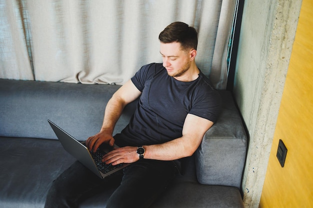 Smiling man in tshirt sitting on sofa typing on netbook working remotely on startup as freelancer looking at laptop and smiling