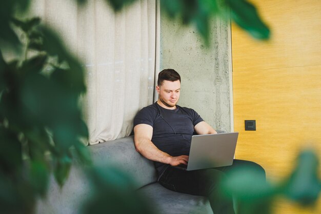 Smiling man in tshirt sitting on sofa typing on netbook working remotely on startup as freelancer looking at laptop and smiling