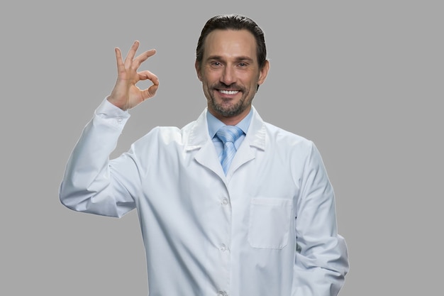 Smiling man in white coat showing okay symbol. Satisfied doctor or scientist is on gray background.