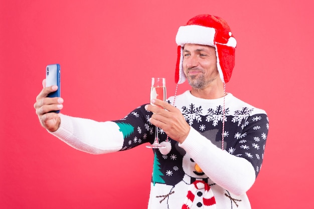 Smiling man in winter sweater and hat making selfie on smartphone with champagne on red background, xmas.