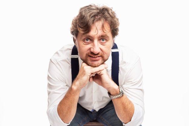 A smiling man with a beard in a white shirt and jeans sits. White wall.