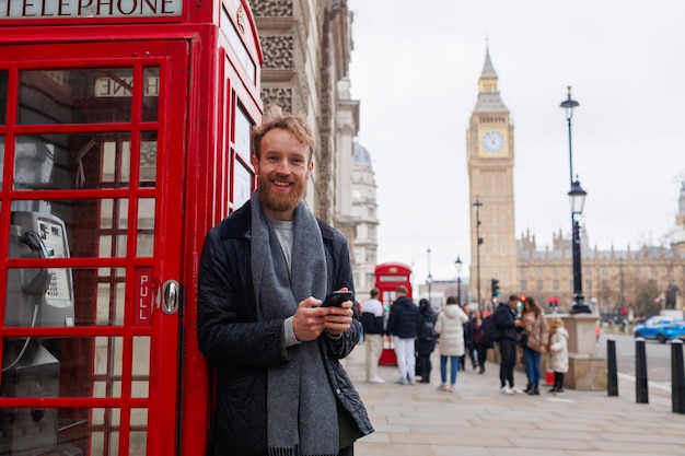 Smiling man with a smartphone in his hands stands near the famous red telephone booth