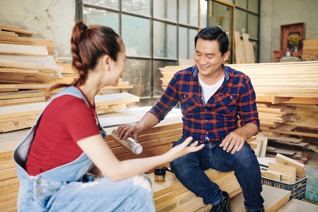 Smiling mature Asian carpenter pouring hot tea from thermos when talking to daughter working in the same workshop