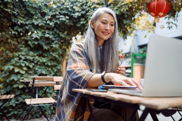 Smiling mature asian woman eats strawberry dessert working on laptop at table outdoors