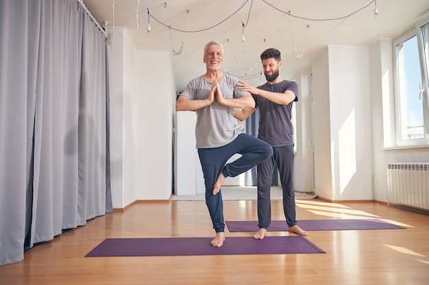 Smiling mature Caucasian beginner yogi doing a tree pose assisted by a qualified yoga instructor