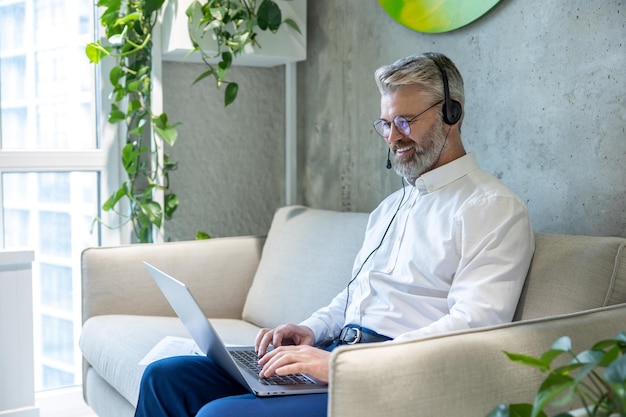 Smiling mature Caucasian businessperson in the headset seated on the sofa typing on the laptop