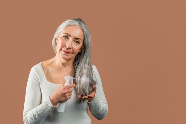 Smiling mature caucasian female with natural beauty applies spray on gray hair looking at camera