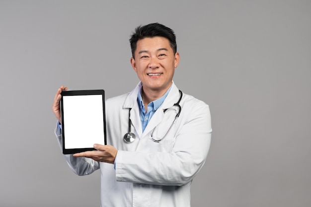 Smiling mature chinese doctor in white coat with stethoscope showing tablet with blank screen