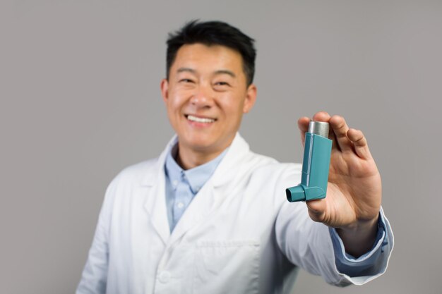 Smiling mature chinese male therapist in white coat showing inhaler for breathing and asthmatic attack