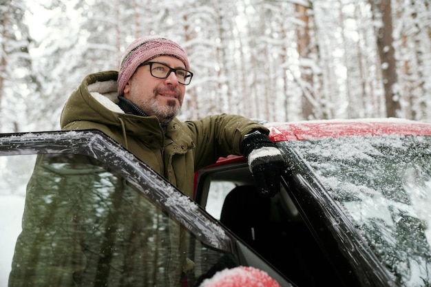 Smiling mature man in eyeglasses and winterwear standing by door of his car