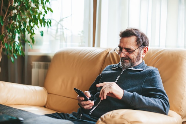 Smiling mature man in glasses sitting on a couch texting or messaging or searching or reading on a modern smartphone Seniors using technology concept