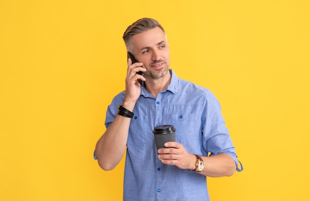 Smiling mature man speaking on phone with coffee cup on yellow background morning