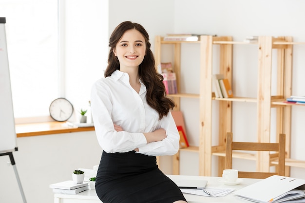 Smiling mature professional businesswoman with arms crossed sitting on the desk in office.