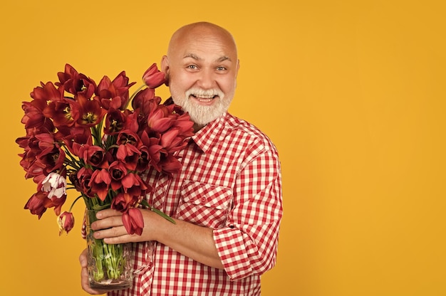 Smiling mature retired man with beard hold spring tulip flowers on yellow background