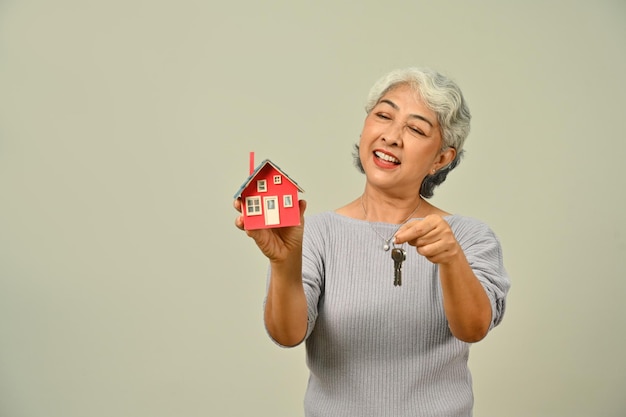 Smiling mature woman holding house model and keys isolated on beige background Real estate building property and insurance