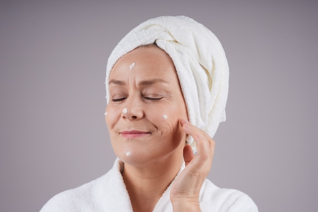 Smiling mature woman with a white towel on her head, applying cream to her face, closing her eyes. Facial care concept. Isolate on gray wall