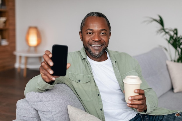 Smiling middle aged african american man in casual with cup of drink takeaway show smartphone with empty screen