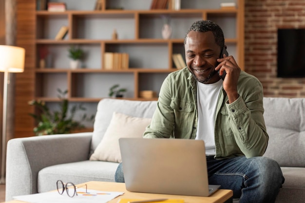 Smiling middle aged black male manager typing on computer calling by phone talk with client