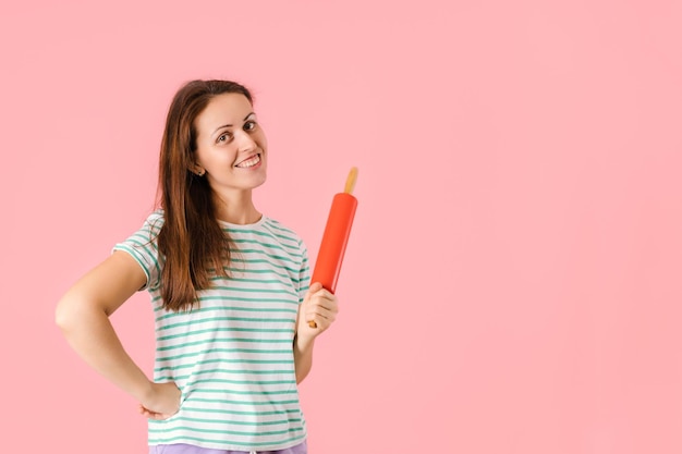 A smiling middle-aged brunette holds a red rolling pin in her hands while standing on a pink background