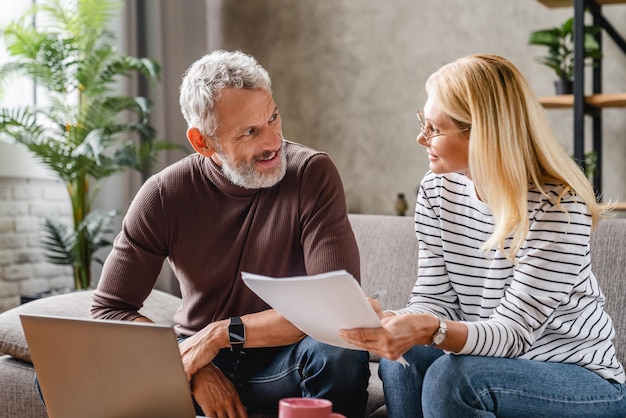 Smiling middle aged couple with laptop studying documents while working on couch at home