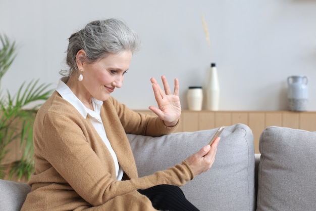 Smiling middle aged woman holding phone, using mobile device apps, looking at screen, while sitting on couch.