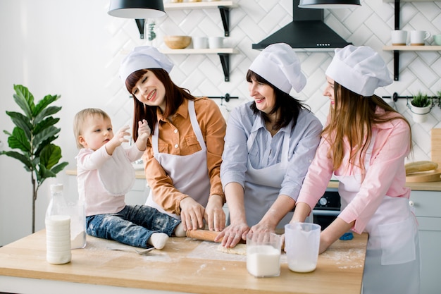 Smiling middle aged woman in kitchen apron rolling out dough and two daughters helping her. Litle baby girl sitting on the table and having fun. Happy women in white aprons baking together